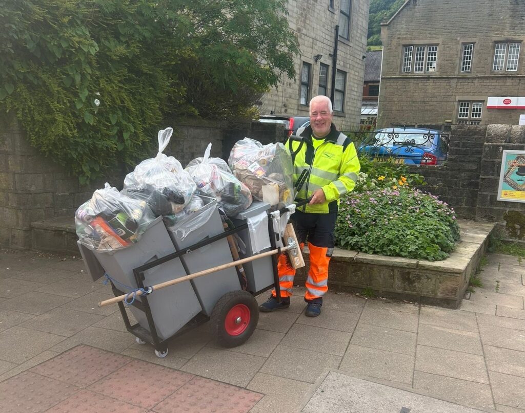 A cleaning operative, wearing a high-visibility jacket and orange trousers, stands next to a barrow filled with bags of rubbish