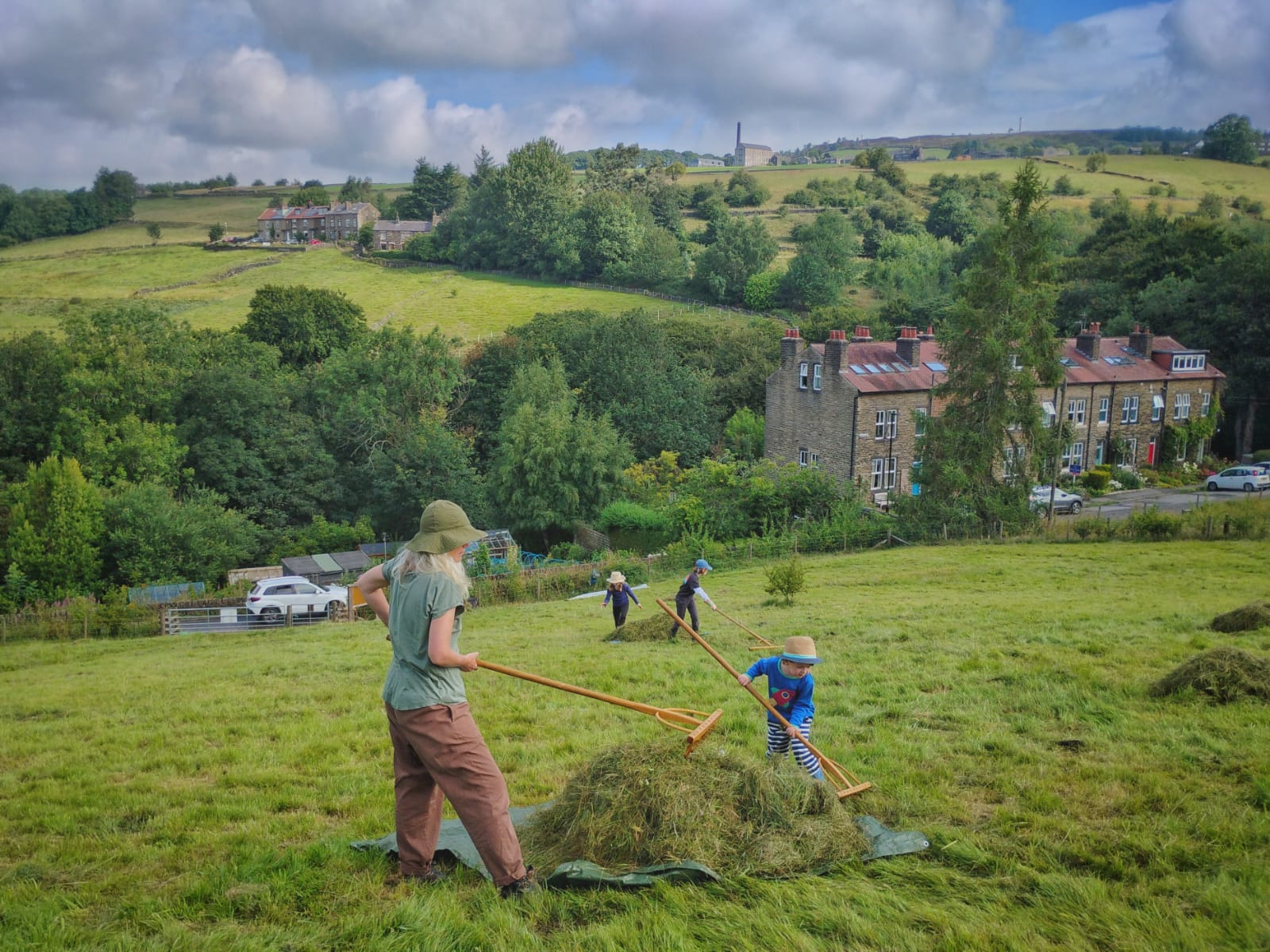 Haytime 2025 adults and children raking hay onto a tarpaulin with Pecket Well mill on the horizon