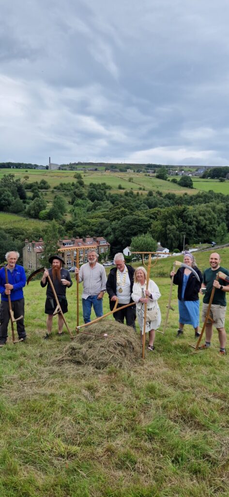 A group of volunteers, members of the Friends of High Hirst Woodmeadow and the Mayor of Hebden Royd Town Council, Cllr. Keith Butterick, stand around a pile of recently mown hay on High Hirst Woodmeadow.