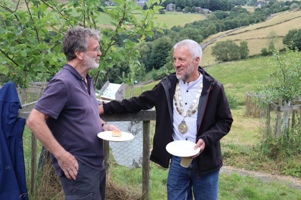 Two people talking to one another in a meadow, with fields in the background.