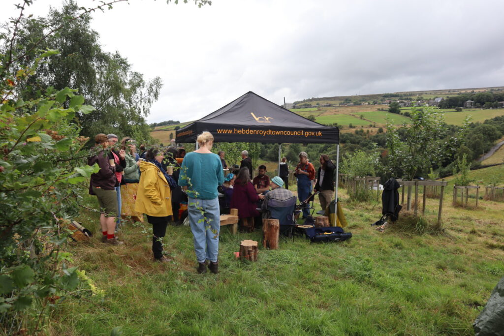 Haytime Tea celebration at High Hirst Woodmeadow in Hebden Bridge. A group of people gather around a gazebo, with a collective of folk musicians playing live music and a buffet is served.
